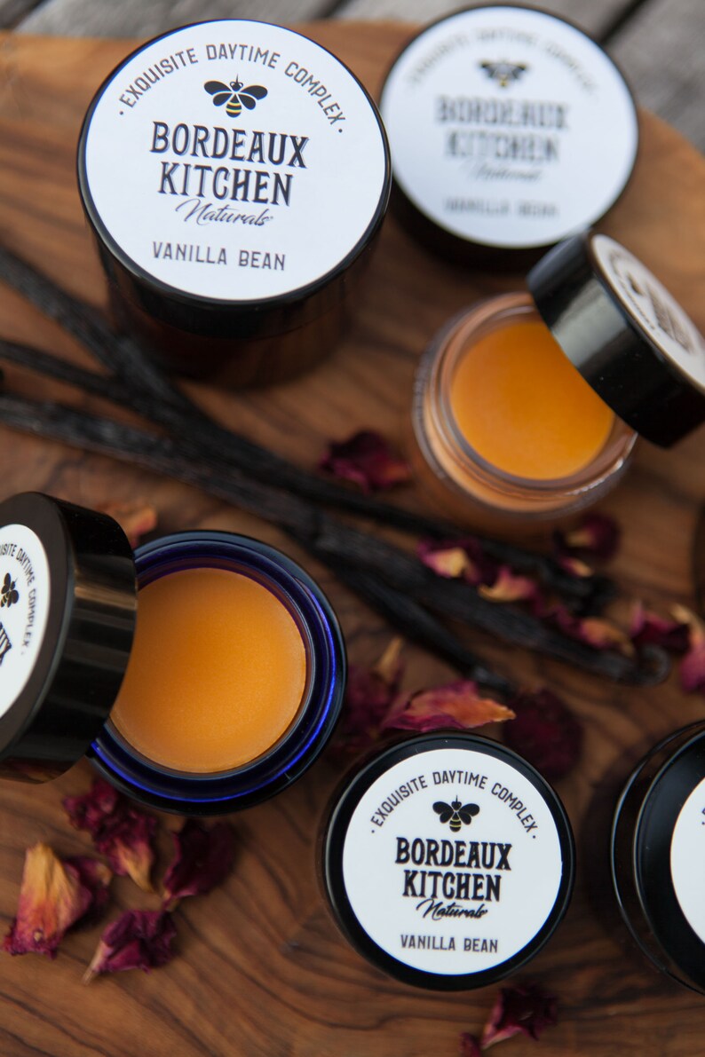 K&ouml;nnte beinhalten: Vier kleine Gl&auml;ser mit Lippenbalsam auf einer Holzoberfl&auml;che mit Vanilleschoten und Rosenbl&uuml;ten. Die Gl&auml;ser sind mit "Bordeaux Kitchen Naturals Vanilla Bean" beschriftet und zeigen ein Bienenlogo.