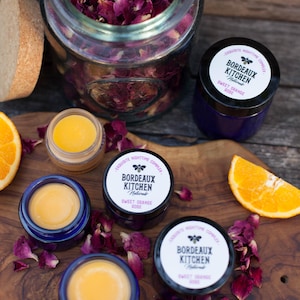 Puede incluir: Tres pequeños frascos de vidrio con tapas azules llenos de un bálsamo amarillo. Los frascos tienen etiquetas que dicen "Bordeaux Kitchen Naturale Sweet Orange Rose" y están rodeados de pétalos de rosa sobre una superficie de madera. Hay un frasco de vidrio más grande lleno de pétalos de rosa secos y una rodaja de naranja sobre la superficie.