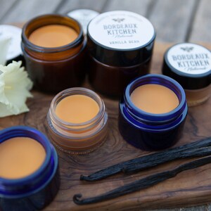 K&ouml;nnte beinhalten: Ein Holzbrett mit f&uuml;nf kleinen Gl&auml;sern Lippenbalsam. Die Gl&auml;ser sind in verschiedenen Farben, darunter blau, braun und klar. Der Lippenbalsam ist hellorange. Auf dem Brett liegen auch zwei Vanilleschoten. Der Text auf den Gl&auml;sern lautet "Bordeaux Kitchen Natural" und "Vanilla Bean".