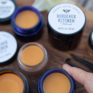 K&ouml;nnte beinhalten: F&uuml;nf kleine Gl&auml;ser Lippenbalsam, vier mit einem durchsichtigen Deckel und einem blauen Rand und eines mit einem schwarzen Deckel. Das Glas mit dem schwarzen Deckel hat ein wei&szlig;es Etikett mit der Aufschrift "Bordeaux Kitchen Naturals Vanilla Bean".