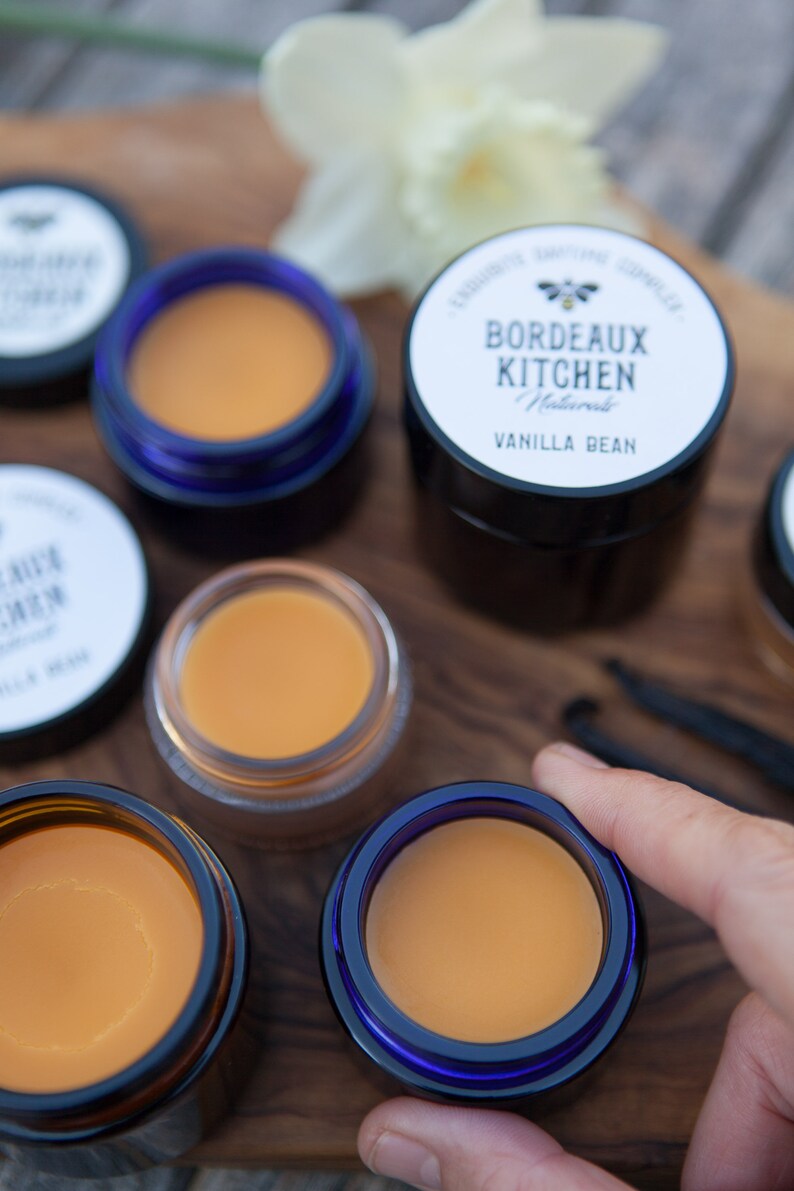 K&ouml;nnte beinhalten: F&uuml;nf kleine Gl&auml;ser Lippenbalsam, vier mit einem durchsichtigen Deckel und einem blauen Rand und eines mit einem schwarzen Deckel. Das Glas mit dem schwarzen Deckel hat ein wei&szlig;es Etikett mit der Aufschrift "Bordeaux Kitchen Naturals Vanilla Bean".