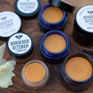 K&ouml;nnte beinhalten: Nahaufnahme von sechs kleinen Gl&auml;sern Lippenbalsam, alle mit dem Label "Bordeaux Kitchen Naturals" und einem Bienenlogo. Die Gl&auml;ser sind auf einer Holzoberfl&auml;che mit einer wei&szlig;en Blume und Vanilleschoten angeordnet.