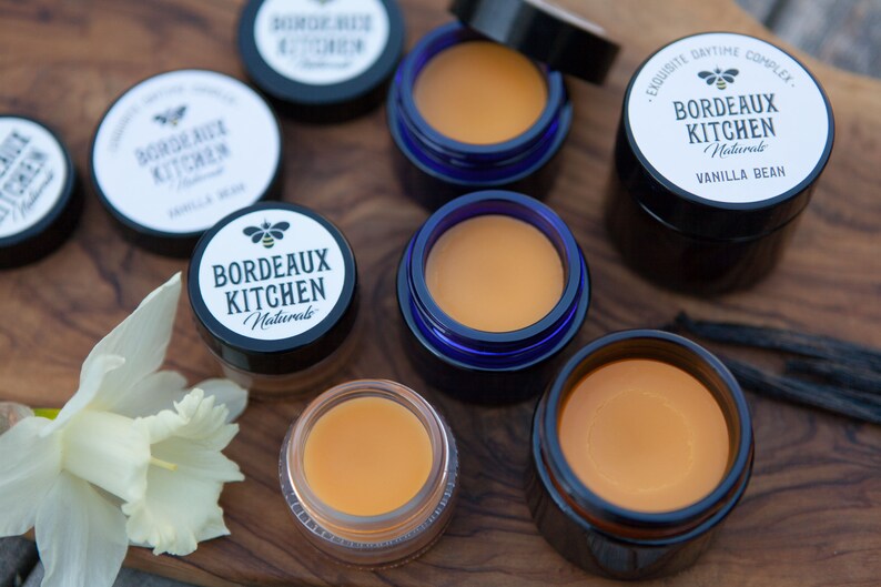 K&ouml;nnte beinhalten: Nahaufnahme von sechs kleinen Gl&auml;sern Lippenbalsam, alle mit dem Label "Bordeaux Kitchen Naturals" und einem Bienenlogo. Die Gl&auml;ser sind auf einer Holzoberfl&auml;che mit einer wei&szlig;en Blume und Vanilleschoten angeordnet.
