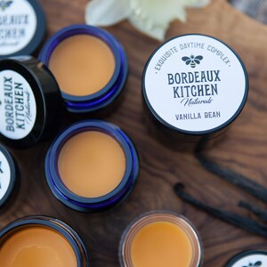 K&ouml;nnte beinhalten: Nahaufnahme von sechs kleinen Gl&auml;sern Lippenbalsam auf einer Holzoberfl&auml;che. Die Gl&auml;ser sind mit "Bordeaux Kitchen Naturals" und "Vanilla Bean" beschriftet.