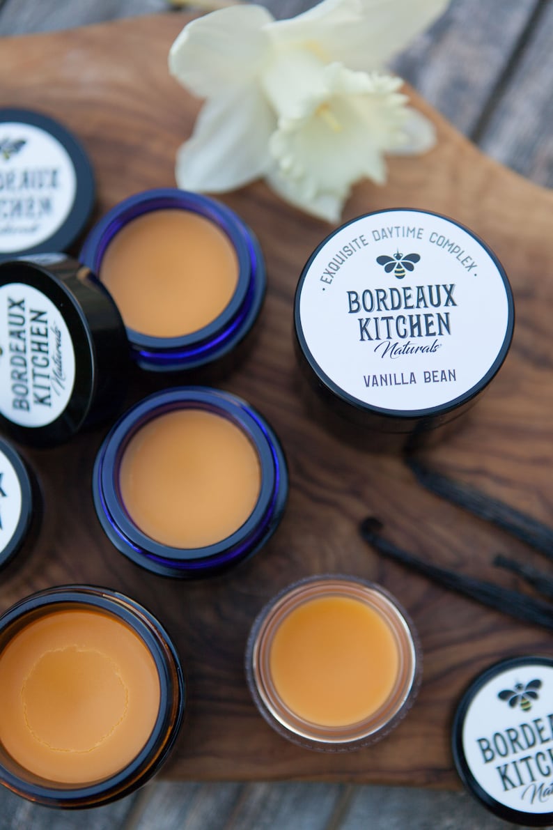 K&ouml;nnte beinhalten: Nahaufnahme von sechs kleinen Gl&auml;sern Lippenbalsam auf einer Holzoberfl&auml;che. Die Gl&auml;ser sind mit "Bordeaux Kitchen Naturals" und "Vanilla Bean" beschriftet.