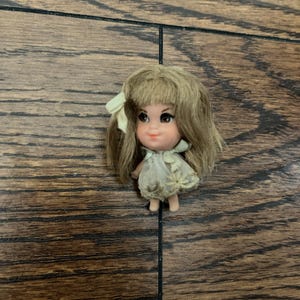 May include: A small vintage doll with blonde hair and a light green bow. The doll has large brown eyes and a light pink face. She is wearing a light-colored dress. The doll is a collectible item.