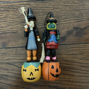 May include: Two wooden Halloween witch figurines. One witch is dressed in black and gray with a straw broom. The other witch is dressed in black, red, and blue with a straw broom. Both witches stand on carved pumpkin bases.