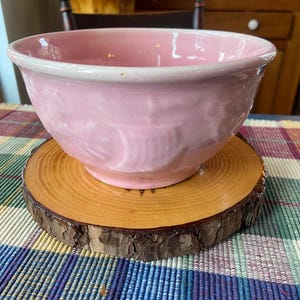 May include: A pink ceramic bowl with a white rim sits on a wooden slice. The bowl has a textured surface and is slightly chipped on the rim.