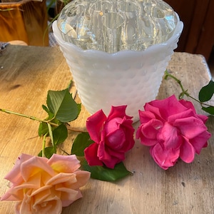 May include: A white milk glass bud vase with a clear glass insert for holding flowers. Three roses, one peach and two pink, are placed on a wooden table next to the vase.