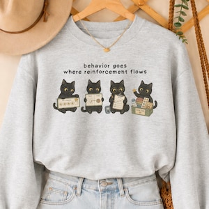 May include: A heather gray crewneck sweatshirt featuring a graphic of four black cats with the text "behavior goes where reinforcement flows". The cats are shown holding various items including a chart, a clipboard, a document, and a box of supplies.