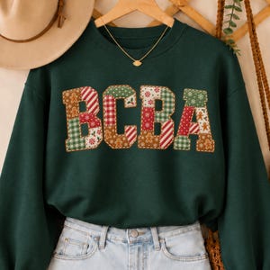 May include: A forest green long-sleeve sweatshirt featuring the letters BCBA in a patchwork design. The letters are filled with various holiday-themed patterns including gingerbread men, red and white stripes, green plaid, and snowflakes.