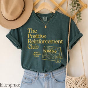 May include: A dark teal short-sleeve t-shirt featuring yellow text that reads The Positive Reinforcement Club, Notice The Good Stuff, and You Did It above a graphic of a star chart with a bucket. A tan hat and a beige woven bag are nearby.