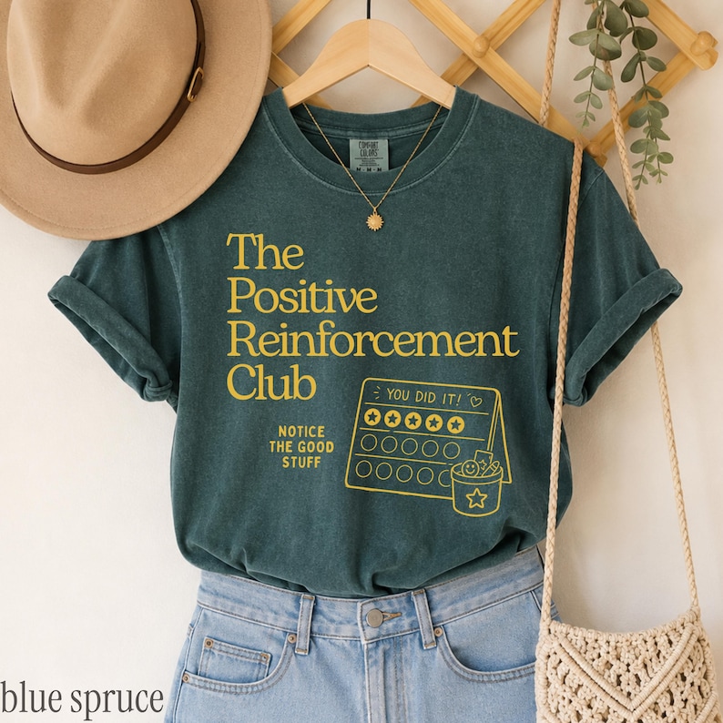 May include: A dark teal short-sleeve t-shirt featuring yellow text that reads The Positive Reinforcement Club, Notice The Good Stuff, and You Did It above a graphic of a star chart with a bucket. A tan hat and a beige woven bag are nearby.