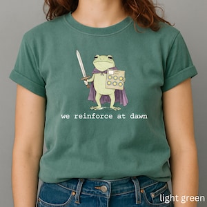 May include: Light green t-shirt featuring a cartoon frog character wearing a purple cape and holding a sword and a star-covered card. The text "we reinforce at dawn" is printed in white below the frog illustration. The shirt is a solid light green color.