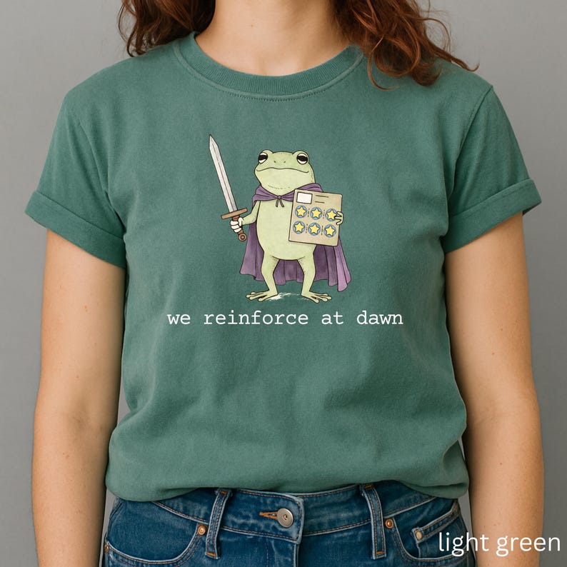 May include: Light green t-shirt featuring a cartoon frog character wearing a purple cape and holding a sword and a star-covered card. The text "we reinforce at dawn" is printed in white below the frog illustration. The shirt is a solid light green color.