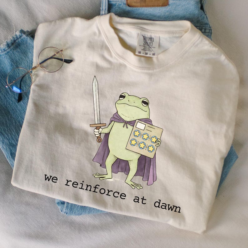May include: Cream-colored t-shirt featuring a cartoon frog warrior with a sword and cape, holding a star-studded card. The text "we reinforce at dawn" is printed at the bottom. The shirt is folded on top of blue jeans.