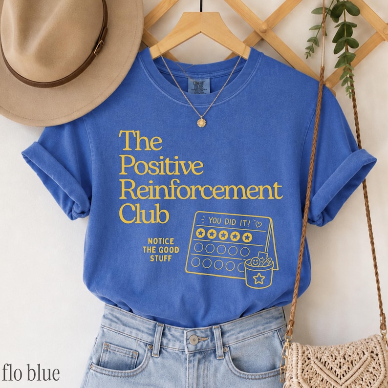 May include: A blue short-sleeve t-shirt featuring yellow text that reads The Positive Reinforcement Club, Notice The Good Stuff, and You Did It, along with a graphic of a reward chart with stars and a bucket icon, displayed on a hanger.