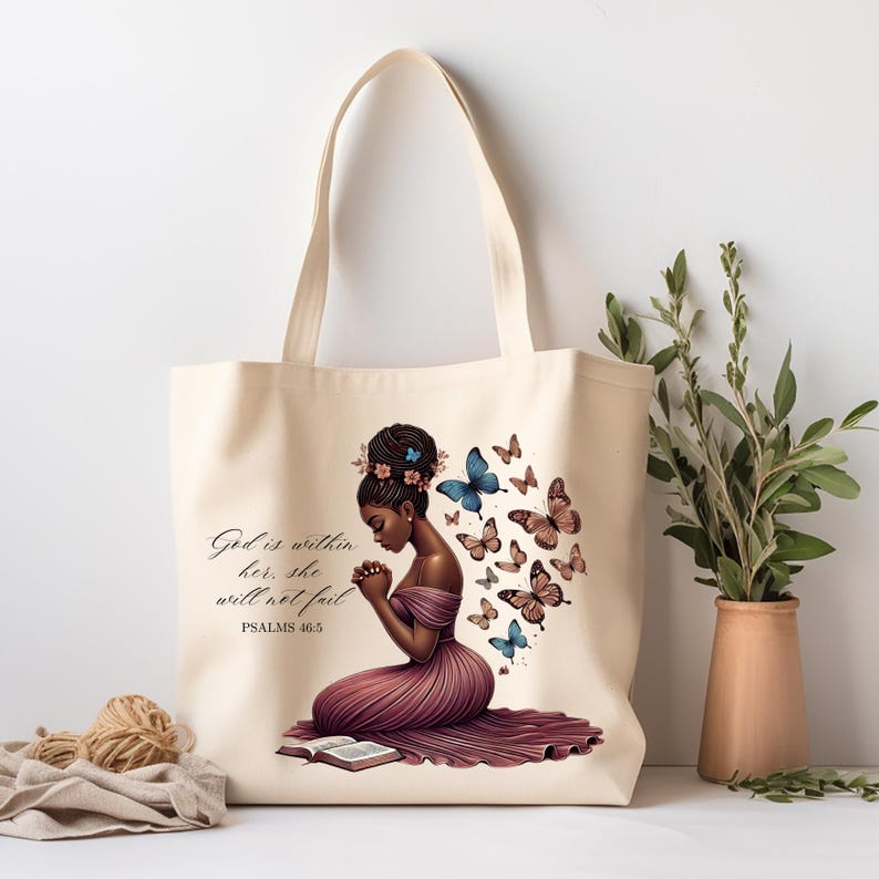 May include: A beige tote bag with a design of a woman kneeling with her hands clasped in prayer. The woman has dark skin and is wearing a pink dress. The text "God is within her, she will not fail. PSALMS 46:5" is printed on the bag. There are butterflies in various colors surrounding the woman.