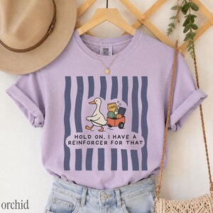 May include: A light purple short-sleeve t-shirt featuring a graphic of a white duck pulling a red wagon filled with items. The text reads, "HOLD ON, I HAVE A REINFORCER FOR THAT." The design is set against a background of vertical dark blue stripes.