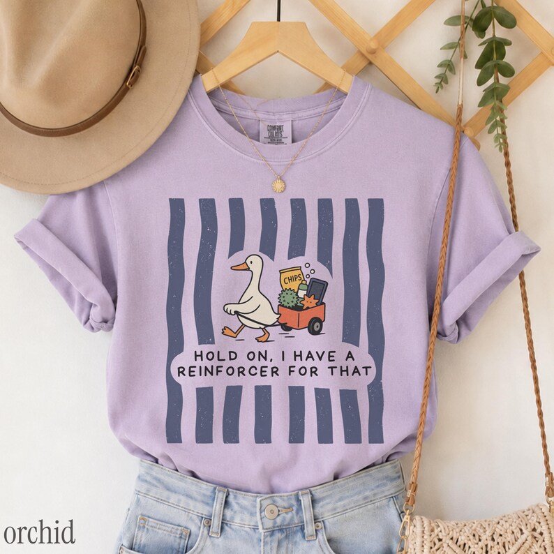 May include: A light purple short-sleeve t-shirt featuring a graphic of a white duck pulling a red wagon filled with items. The text reads, "HOLD ON, I HAVE A REINFORCER FOR THAT." The design is set against a background of vertical dark blue stripes.