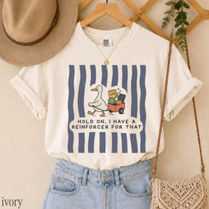May include: An ivory t-shirt featuring a graphic of a white duck pulling a red wagon filled with items, set against vertical navy blue stripes. The text reads, "HOLD ON, I HAVE A REINFORCER FOR THAT." The shirt is paired with light blue denim jeans.