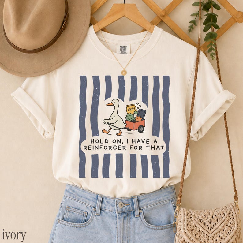 May include: An ivory t-shirt featuring a graphic of a white duck pulling a red wagon filled with items, set against vertical navy blue stripes. The text reads, "HOLD ON, I HAVE A REINFORCER FOR THAT." The shirt is paired with light blue denim jeans.