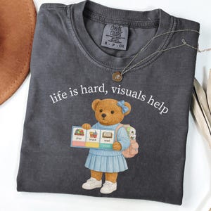 May include: Dark gray t-shirt with the text "life is hard, visuals help." The shirt features a cartoon bear holding a sign with the words "play," "snack," and "read." The bear is wearing a blue dress and carrying a backpack. A necklace is draped across the shirt.