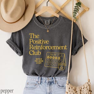 May include: A charcoal gray short-sleeve t-shirt featuring yellow text that reads The Positive Reinforcement Club and Notice The Good Stuff, alongside a yellow graphic of a reward chart with the text You Did It and a star-adorned bucket.