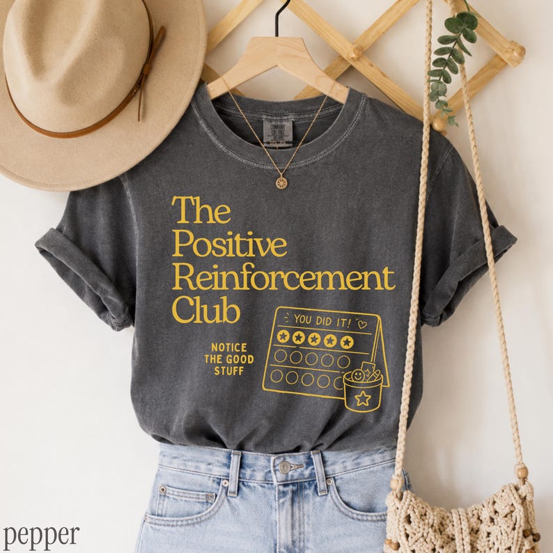 May include: A charcoal gray short-sleeve t-shirt featuring yellow text that reads The Positive Reinforcement Club and Notice The Good Stuff, alongside a yellow graphic of a reward chart with the text You Did It and a star-adorned bucket.