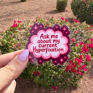 May include: A sticker with the text "Ask me about my current Hyperfixation" in a cloud-shaped design. The sticker has a pink and burgundy color scheme with star accents. The sticker is held in front of a background of red flowers and green bushes.
