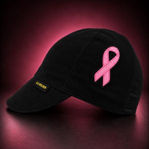 May include: A black welder cap featuring a prominent pink ribbon patch on the side. The cap has a short, rounded brim and a small rectangular black tag on the brim with the yellow text U.S. WELDER stitched onto it. The fabric has visible seams.