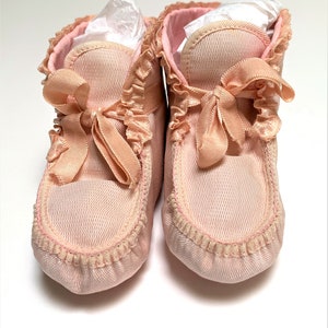 May include: A pair of soft, light pink fabric booties featuring a textured mesh-like exterior, ruffled trim along the ankle opening, and a matching satin ribbon tie closure at the front. The interior is lined with a smooth, solid pink fabric.