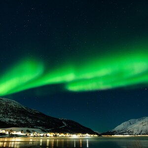 Puede incluir: Un impresionante paisaje nocturno con la Aurora Boreal. Luces verdes vibrantes bailan en el cielo oscuro sobre montañas cubiertas de nieve y una tranquila masa de agua. La escena está iluminada por las luces de un pequeño pueblo.