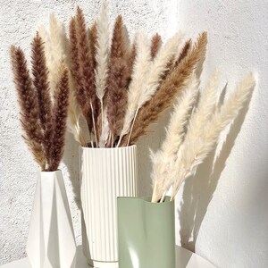 May include: Three vases with dried pampas grass. The vases are white, green, and white with vertical lines. The pampas grass is brown and white.