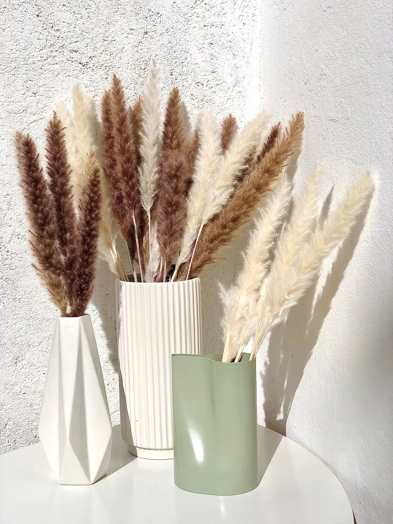 May include: Three vases with dried pampas grass. The vases are white, green, and white with vertical lines. The pampas grass is brown and white.