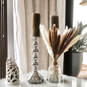 May include: Two brown candles on silver candle holders and a glass vase with dried pampas grass on a white table.