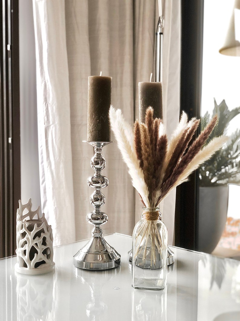 May include: Two brown candles on silver candle holders and a glass vase with dried pampas grass on a white table.