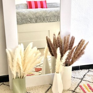 May include: Three white ceramic vases with dried pampas grass in them. The vases are on a white and cream patterned rug. The rug has pink, yellow, and black accents.