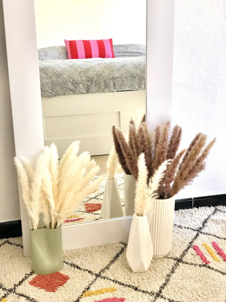 May include: Three white ceramic vases with dried pampas grass in them. The vases are on a white and cream patterned rug. The rug has pink, yellow, and black accents.