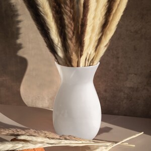 May include: A white ceramic vase filled with dried pampas grass in shades of brown and cream. The vase is sitting on a light brown surface.