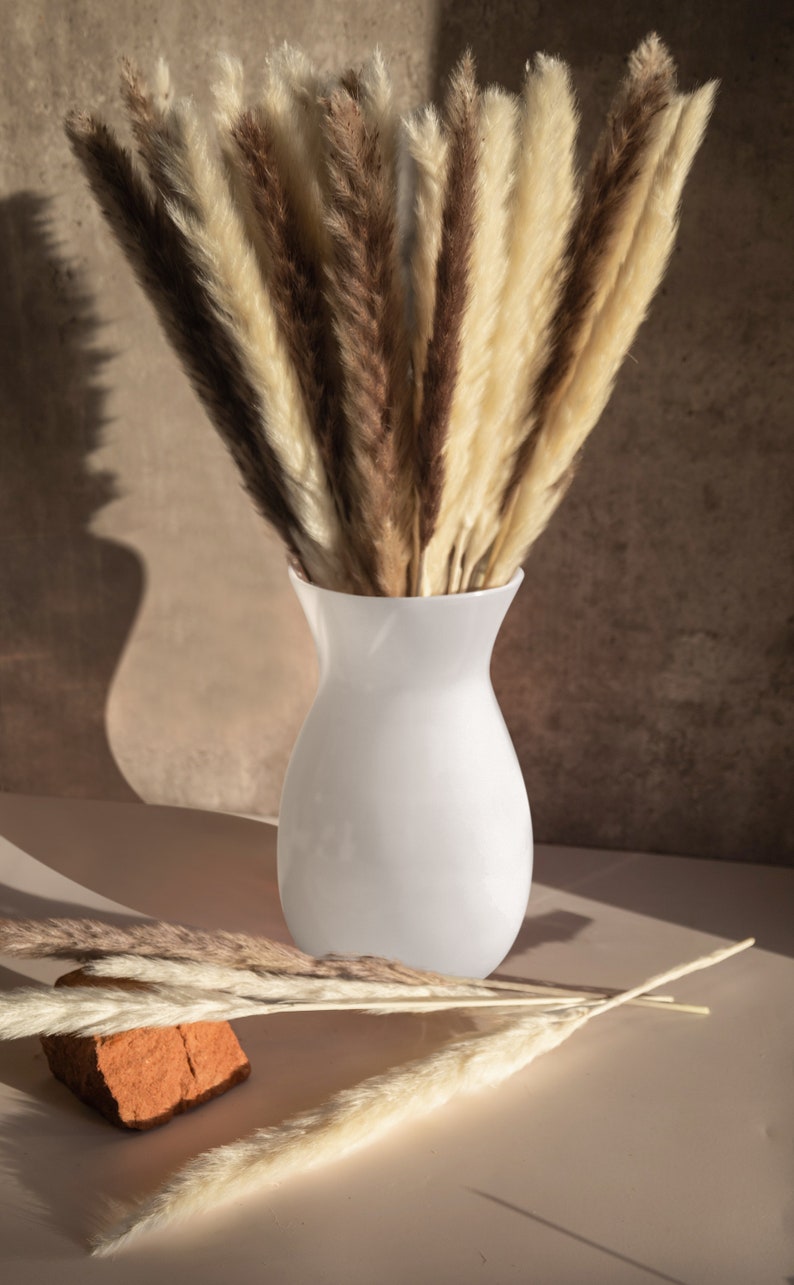 May include: A white ceramic vase filled with dried pampas grass in shades of brown and cream. The vase is sitting on a light brown surface.
