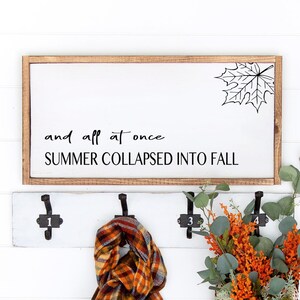 May include: A white wooden sign with black text that reads "and all at once SUMMER COLLAPSED INTO FALL". A black and white outline of a maple leaf is in the top right corner of the sign. The sign is hanging on a white wall with a brown wicker basket, a plaid scarf, and a small wicker basket with orange flowers.