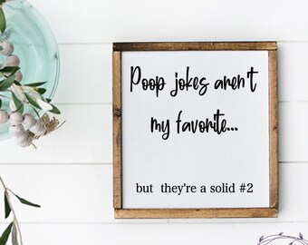 May include: A white wooden sign with brown trim, featuring the text "Poop jokes aren't my favorite... but they're a solid #2". The sign is hanging on a white wood plank wall.