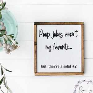 Puede incluir: Un letrero de madera blanco con moldura marrón, con el texto "Poop jokes aren't my favorite... but they're a solid #2". El letrero está colgado en una pared de tablones de madera blanca.