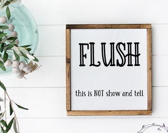 May include: A white wooden sign with a brown frame, featuring the word "FLUSH" in black text. The sign also includes the text "this is NOT show and tell".