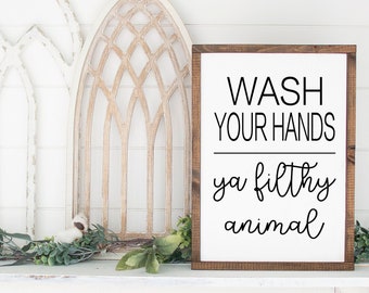 May include: A wooden sign with the text "WASH YOUR HANDS ya filthy animal" in black lettering on a white background. The sign is leaning against a white wall with two decorative wooden arches behind it.