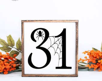 May include: A white wooden sign with a black silhouette of the number 31. A spider is on the web that is behind the number 31.