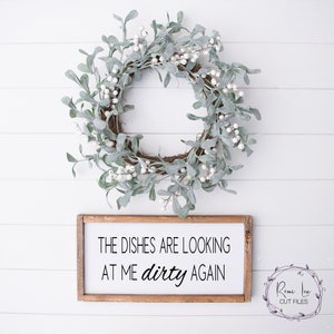 Puede incluir: Un letrero de madera con el texto "The dishes are looking at me dirty again". El letrero está colgado en una pared blanca con una corona blanca y verde encima.