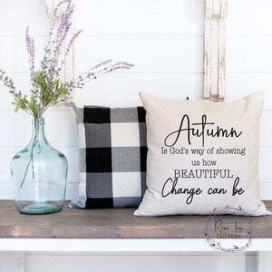 May include: A beige throw pillow with the text "Autumn is God's way of showing us how BEAUTIFUL Change can be" in black script font. The pillow is sitting on a wooden bench with a black and white checkered pillow behind it.