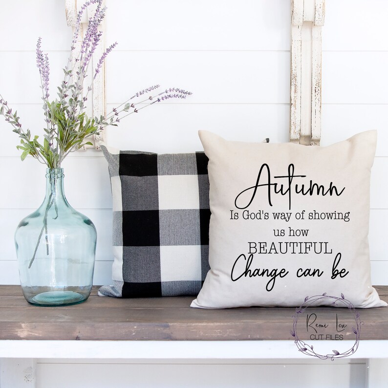May include: A beige throw pillow with the text "Autumn is God's way of showing us how BEAUTIFUL Change can be" in black script font. The pillow is sitting on a wooden bench with a black and white checkered pillow behind it.
